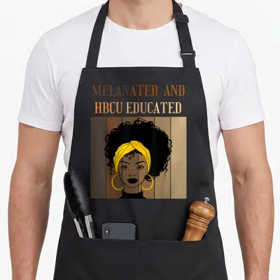 Melanated historical aprons - MELANATED AND HBCU EDUCATED text above woman portrait with yellow headwrap on brown tan — shown on a black cotton canvas apron against a white background