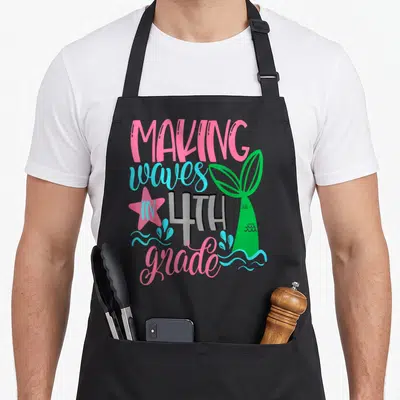 Mermaid making aprons - Green mermaid tail next to stacked pink and cyan text reading MAKING waves IN 4TH grade — shown on a black cotton canvas apron against a white background