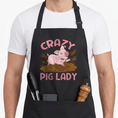 Pig local farm apron - Pink cartoon pig in brown mud with arched CRAZY and straight PIG LADY text — shown on a black cotton canvas apron against a white background