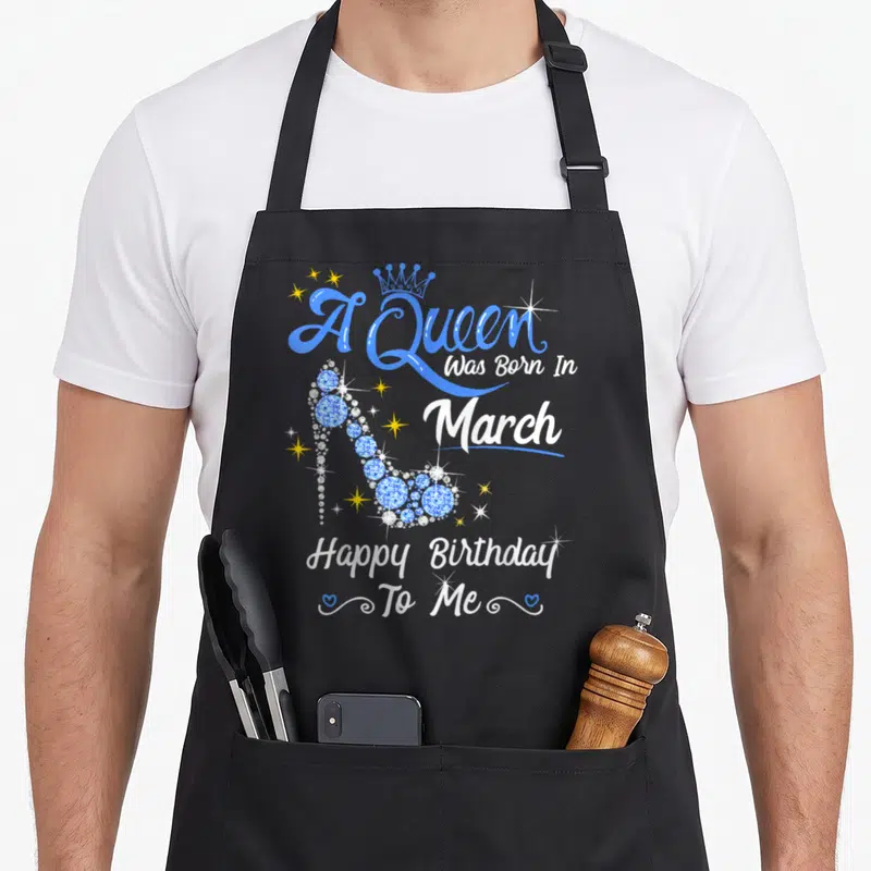 Vintage march baking queen apron - Blue high heel shoe with A Queen Was Born In March text and hearts — shown on a black cotton canvas apron against a white background