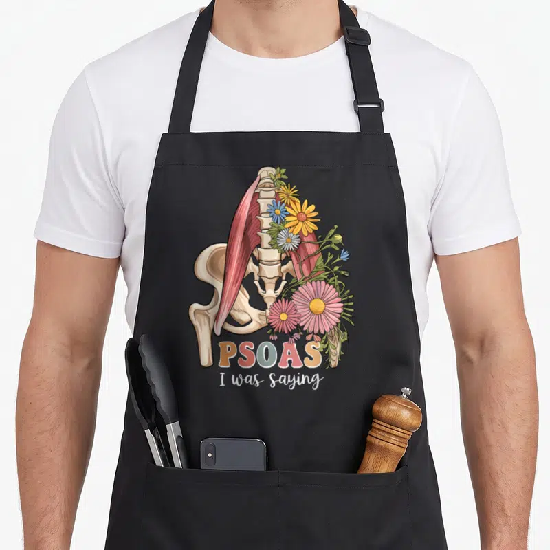 Psoas massage therapy apron - Anatomical psoas muscle spine pelvis with flowers and PSOAS I was saying text — shown on a black cotton canvas apron against a white background