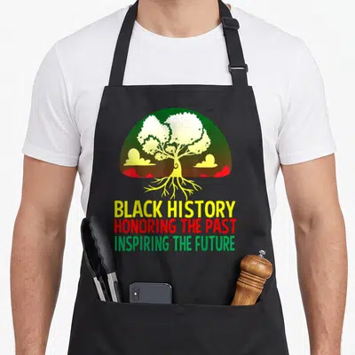Educator matter teacher apron - Yellow tree with roots inside green semi-circle above three lines of text reading Black — shown on a black cotton canvas apron against a white background