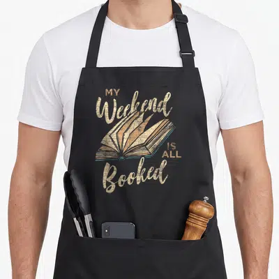 Booked girls weekend apron - Open book illustration with MY Weekend IS ALL Booked text in beige distressed font — shown on a black cotton canvas apron against a white background