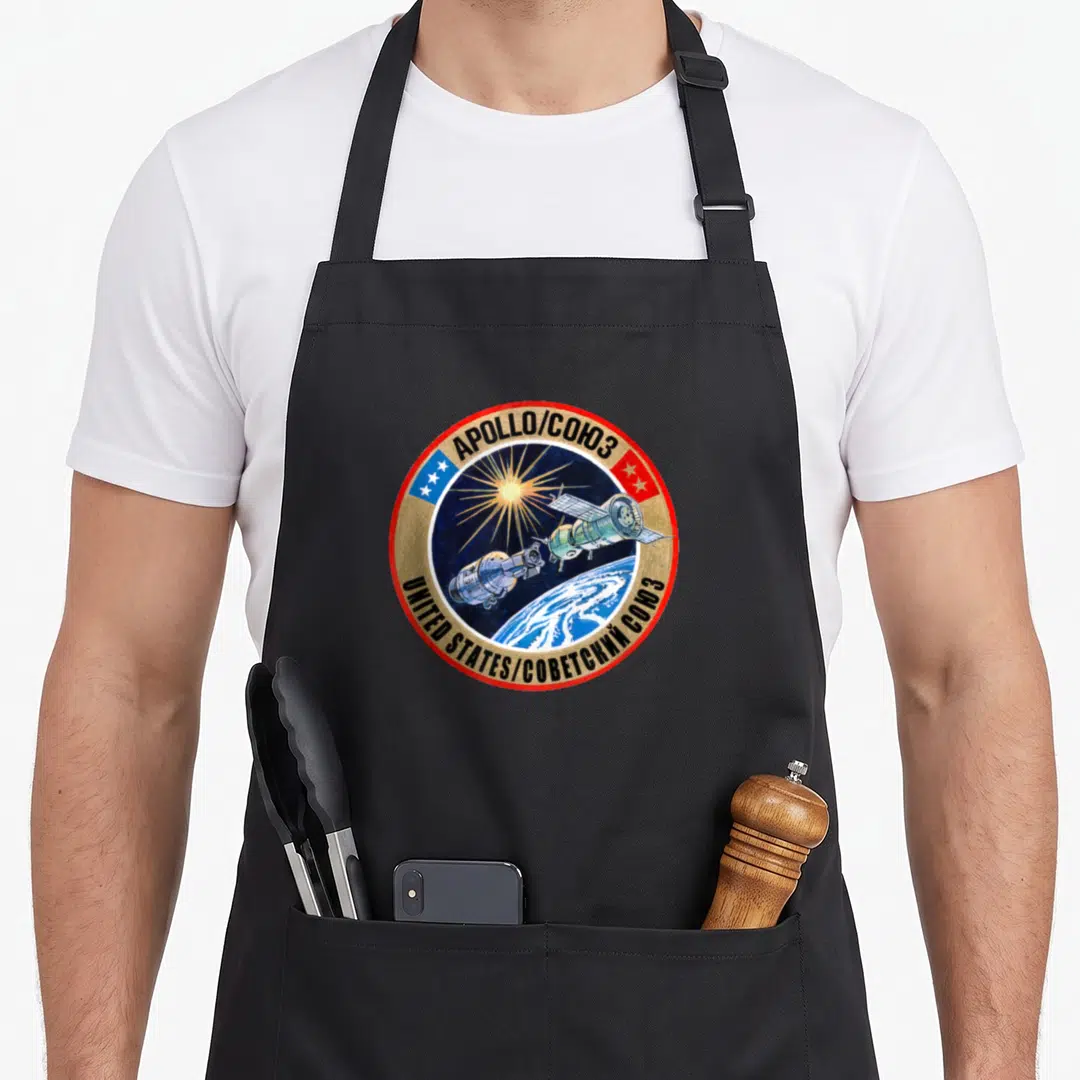 Rendezvous apollo apron - Circular Apollo-Soyuz mission patch with two docked spacecraft and bilingual text — shown on a black cotton canvas apron against a white background