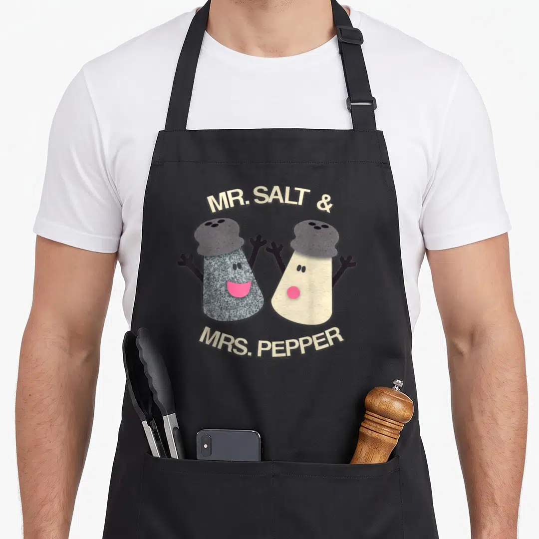 Blue mr mrs aprons - MR. SALT & MRS. PEPPER cartoon shakers with pink mouths on black fabric — shown on a black cotton canvas apron against a white background