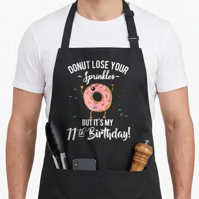 11th donut apron - Pink cartoon donut winking with scattered sprinkles and birthday quote text — shown on a black cotton canvas apron against a white background