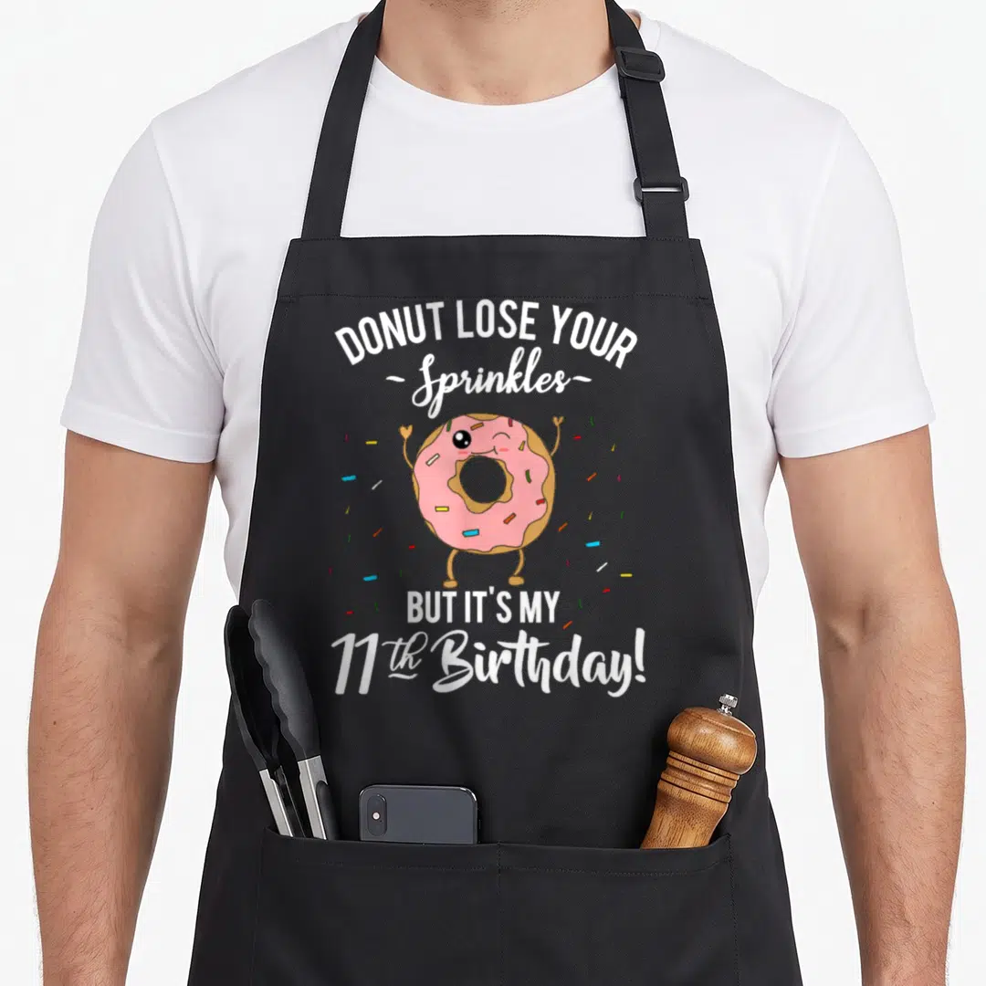 11th donut apron - Pink cartoon donut winking with scattered sprinkles and birthday quote text — shown on a black cotton canvas apron against a white background