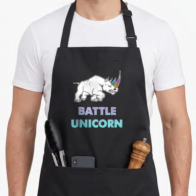 Battle rainbow apron - Cartoon rhinoceros with rainbow horn above BATTLE UNICORN text in purple and cyan — shown on a black cotton canvas apron against a white background