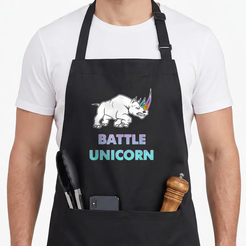 Battle rainbow apron - Cartoon rhinoceros with rainbow horn above BATTLE UNICORN text in purple and cyan — shown on a black cotton canvas apron against a white background