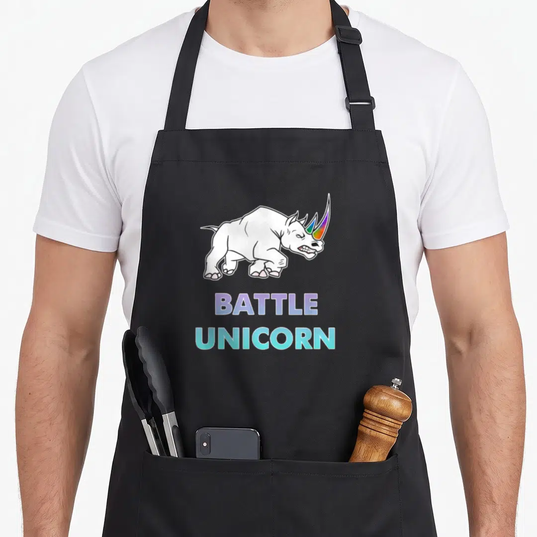Battle rainbow apron - Cartoon rhinoceros with rainbow horn above BATTLE UNICORN text in purple and cyan — shown on a black cotton canvas apron against a white background