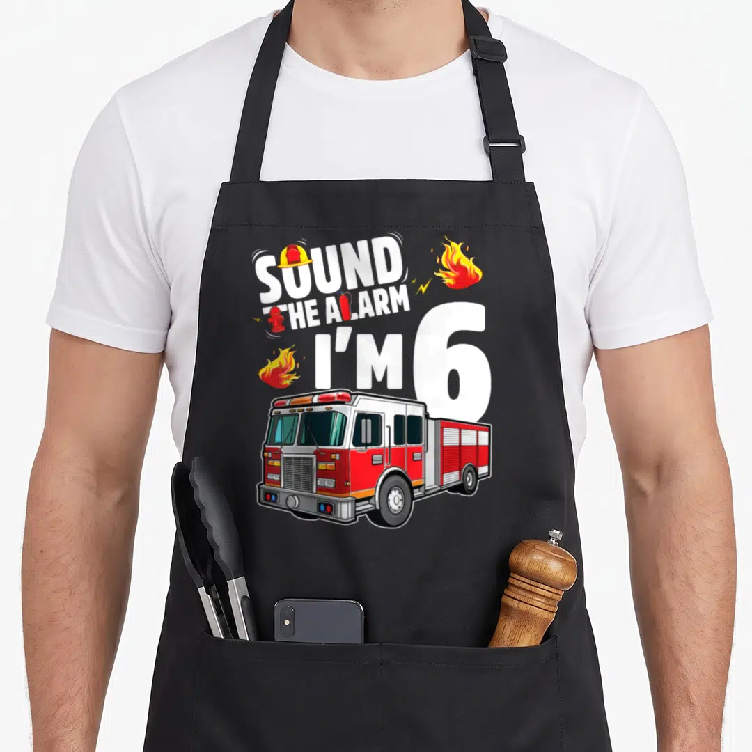 Fire 6th truck apron - SOUND THE ALARM I'M 6 text with fire truck, flames, helmet, hydrant, and extinguisher in red — shown on a black cotton canvas apron against a white background