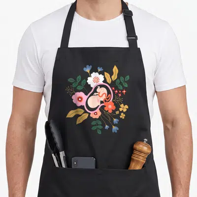 Midwifery floral apron - Fetus in pink womb surrounded by circular floral arrangement in pink green yellow and blue — shown on a black cotton canvas apron against a white background