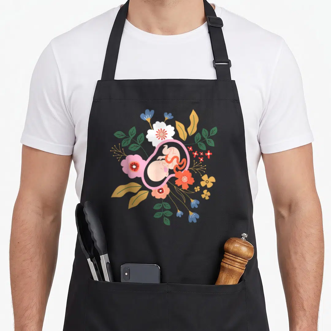 Midwifery floral apron - Fetus in pink womb surrounded by circular floral arrangement in pink green yellow and blue — shown on a black cotton canvas apron against a white background