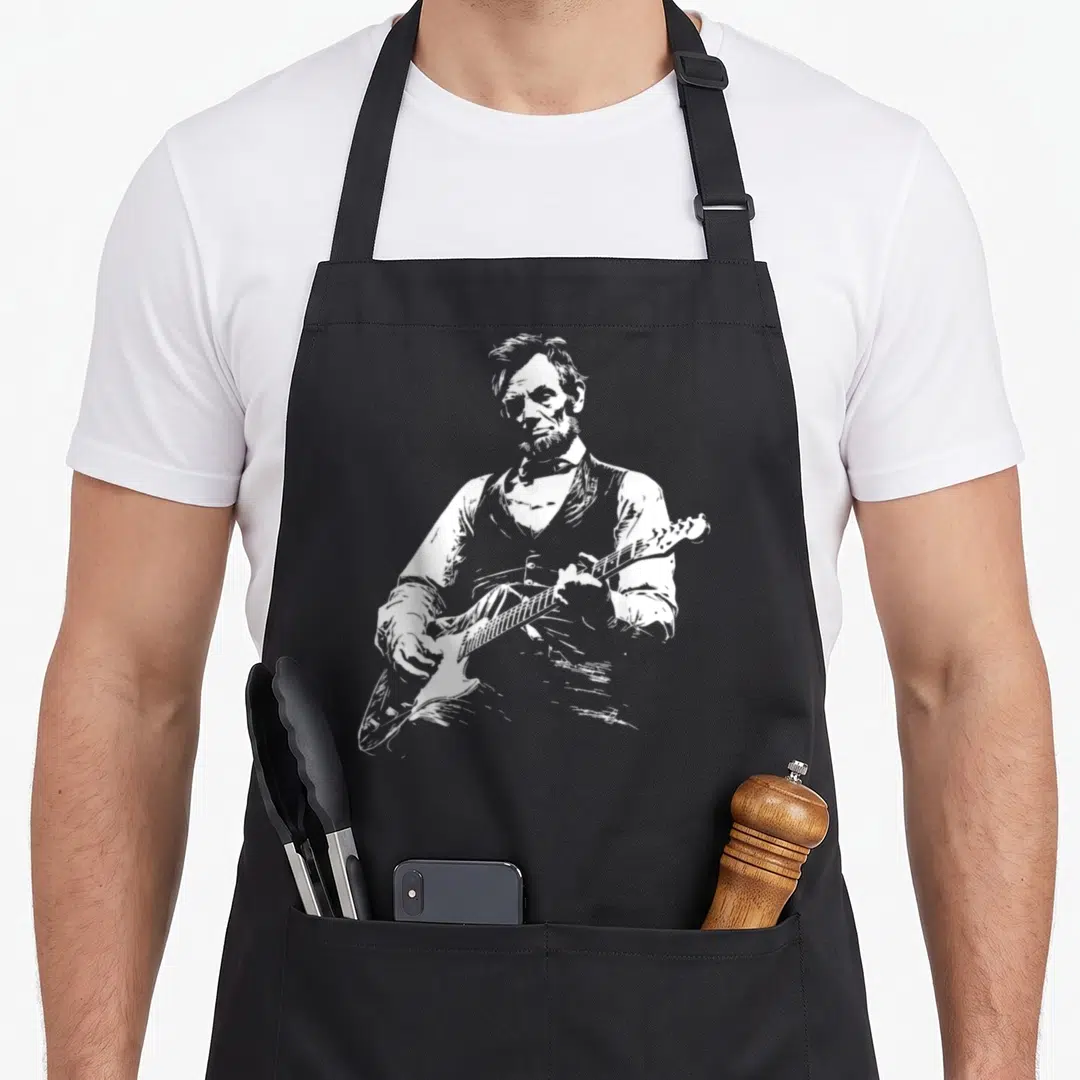 President playing guitar apron - White line-art sketch of Abraham Lincoln playing an electric guitar on dark fabric — shown on a black cotton canvas apron against a white background