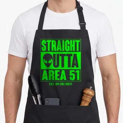Straight funny believer apron - Green bordered text Straight Outta Area 51 with alien head replacing the O — shown on a black cotton canvas apron against a white background