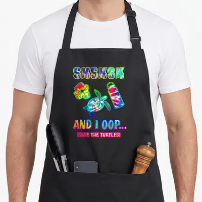 Sksksk tie dye apron - Cartoon turtle, scrunchie, and bottle in tie dye colors with sksksk text — shown on a black cotton canvas apron against a white background