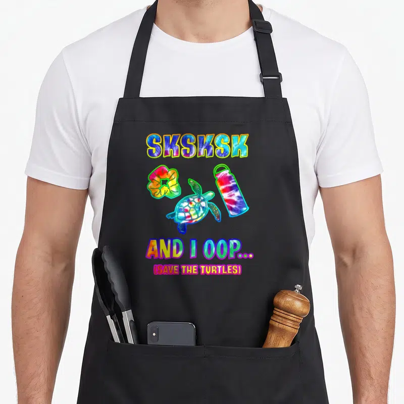 Sksksk tie dye apron - Cartoon turtle, scrunchie, and bottle in tie dye colors with sksksk text — shown on a black cotton canvas apron against a white background