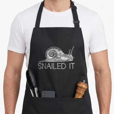 Snailed snail apron - White line art snail above SNAILED IT text on dark background — shown on a black cotton canvas apron against a white background