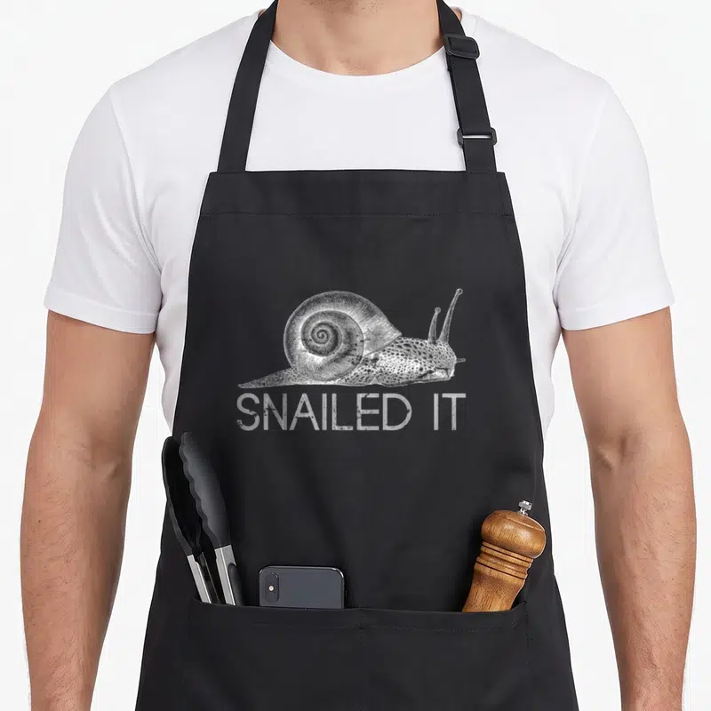 Snailed snail apron - White line art snail above SNAILED IT text on dark background — shown on a black cotton canvas apron against a white background