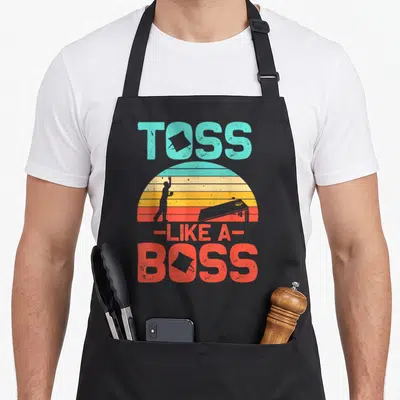 Cornhole boss apron - Retro TOSS LIKE A BOSS text with striped sunset and silhouetted cornhole player tossing bean bag — shown on a black cotton canvas apron against a white background