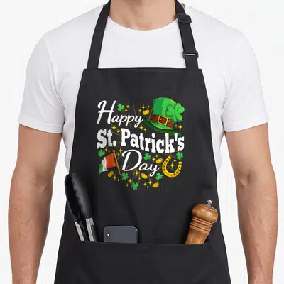 Patrick shamrock irish aprons - Leprechaun hat, flag, horseshoe, coins, and shamrocks surrounding stacked Happy St — shown on a black cotton canvas apron against a white background