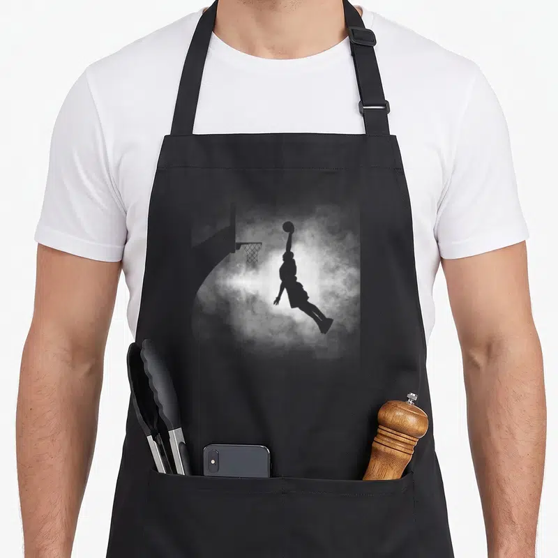 Basketball basbo apron - Grey silhouette of a basketball player jumping toward a hoop against swirling white smoke — shown on a black cotton canvas apron against a white background