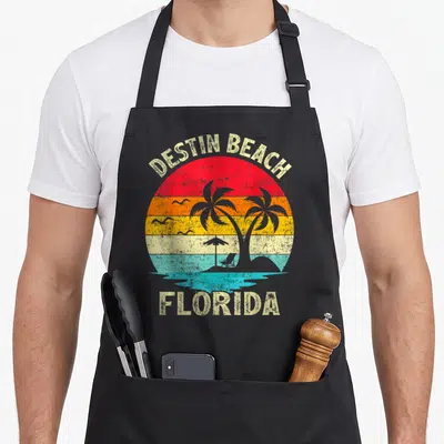 Vacation florida family aprons - Retro Destin Beach Florida sunset with palm trees, beach chair, and multi-colored bands — shown on a black cotton canvas apron against a white background
