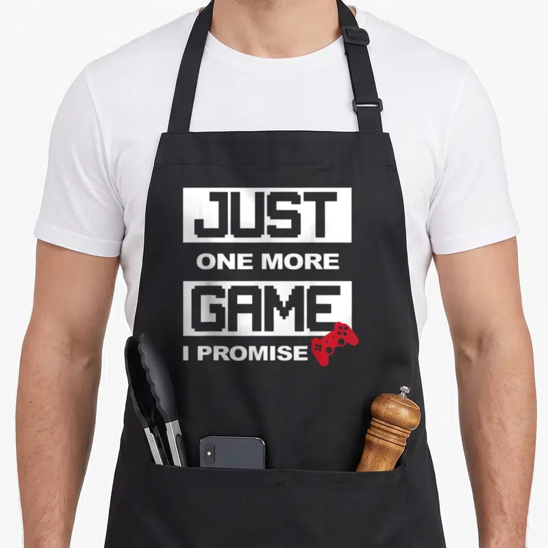 Promise game aprons - Stacked text JUST ONE MORE GAME I PROMISE with red game controller icon — shown on a black cotton canvas apron against a white background