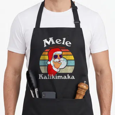 Kalikimaka christmas santa apron - Mele Kalikimaka text above Santa with sunglasses in front of striped sun background — shown on a black cotton canvas apron against a white background