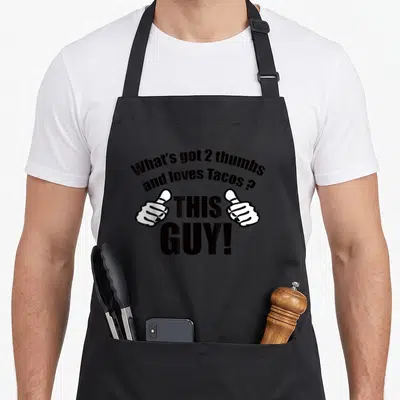Thumb tuesday taco apron - Two white thumbs flanking THIS GUY text with a taco pun above — shown on a black cotton canvas apron against a white background