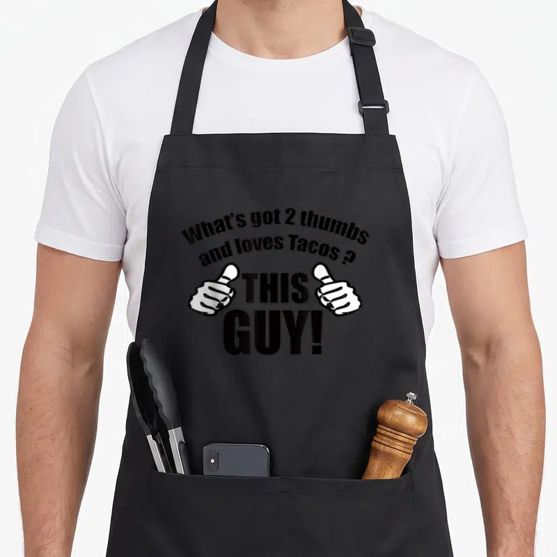 Thumb tuesday taco apron - Two white thumbs flanking THIS GUY text with a taco pun above — shown on a black cotton canvas apron against a white background