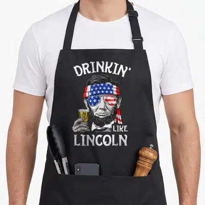 Abraham 4th of july apron - Distressed Lincoln portrait with flag bandana, sunglasses, beer glass, and arched DRINKIN' — shown on a black cotton canvas apron against a white background