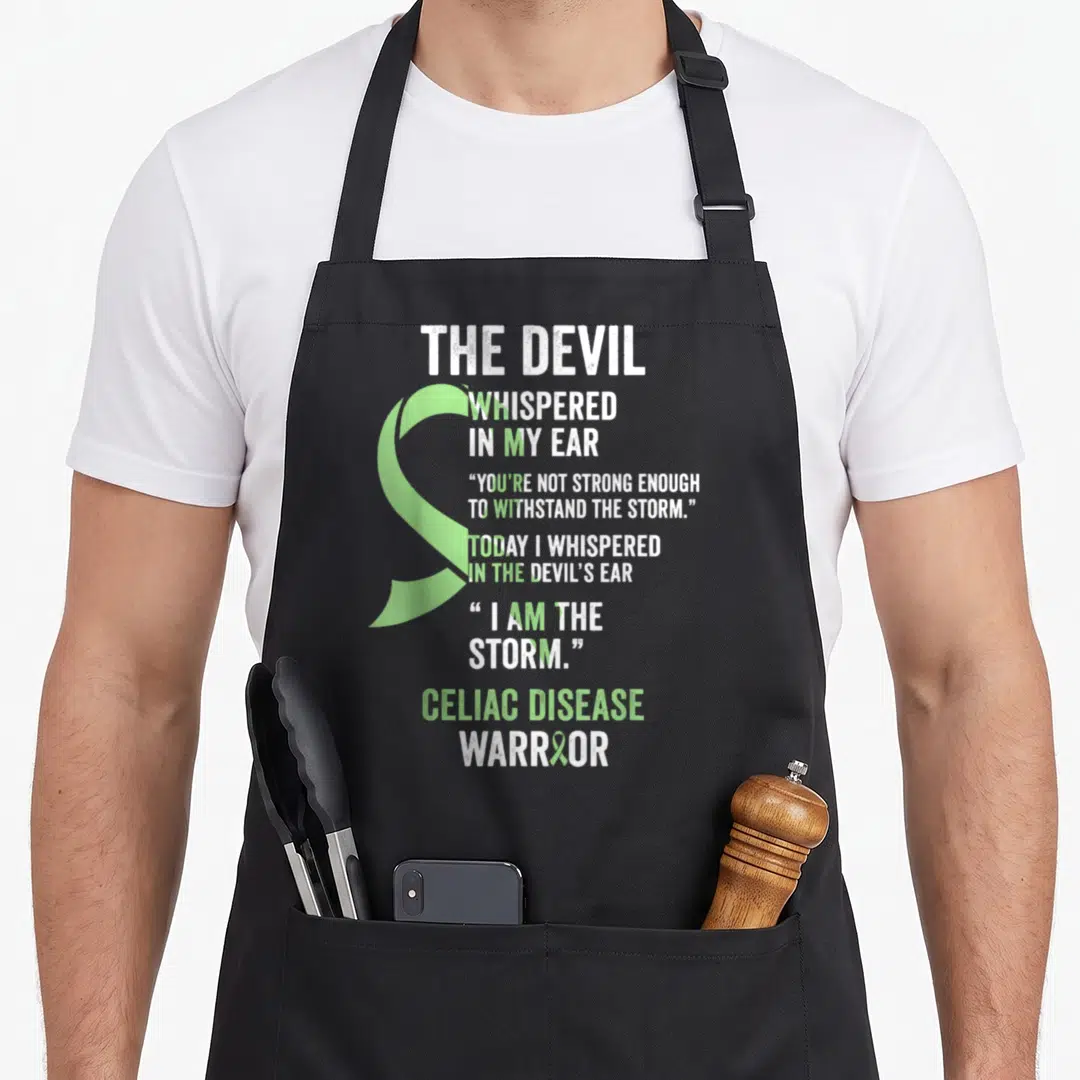 Celiac devil's apron - Light green ribbon overlapping white text reading The Devil Whispered in My Ear Celiac Disease — shown on a black cotton canvas apron against a white background