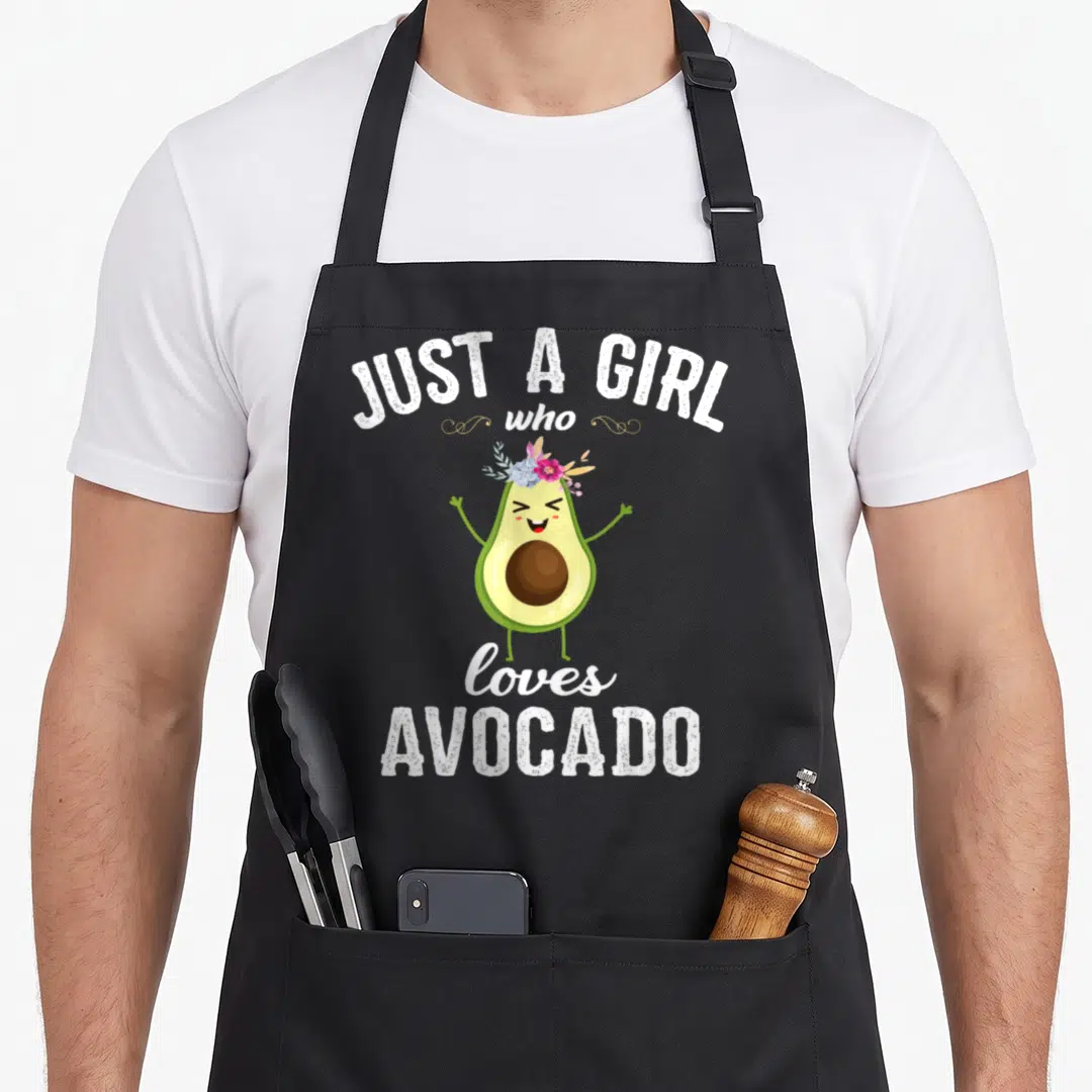Cute avocado apron - Cartoon avocado with pink flower crown and white text Just A Girl Who Loves Avocado — shown on a black cotton canvas apron against a white background