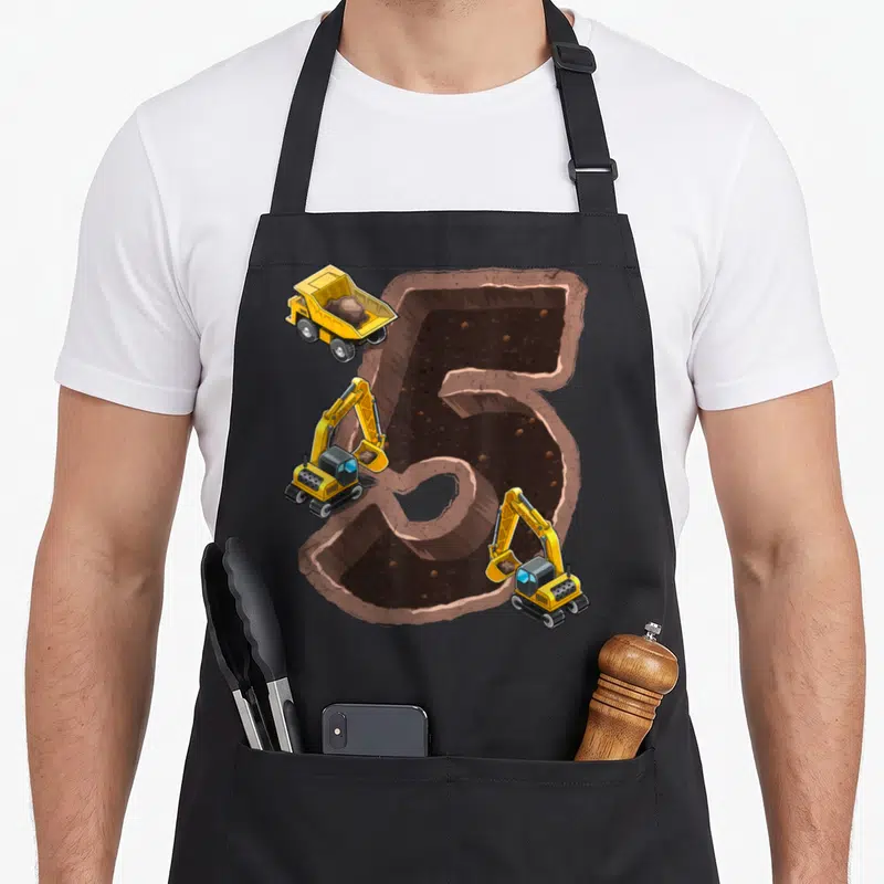 5th birthday vehicle apron - Cartoon number 5 excavation hole with yellow dump truck and two excavators on brown dirt — shown on a black cotton canvas apron against a white background