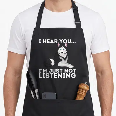 Hear husky apron - Cartoon husky with I HEAR YOU I'M JUST NOT LISTENING text — shown on a black cotton canvas apron against a white background