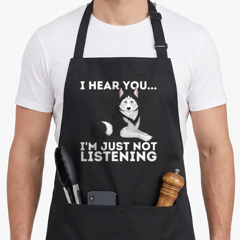 Hear husky apron - Cartoon husky with I HEAR YOU I'M JUST NOT LISTENING text — shown on a black cotton canvas apron against a white background
