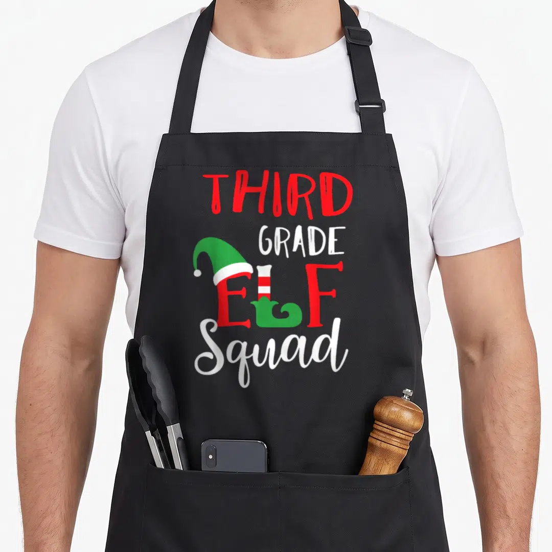 Third christmas elf apron - Stacked THIRD GRADE ELF Squad text with elf hat and shoe in red white green — shown on a black cotton canvas apron against a white background