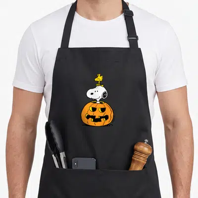 Woodstock pumpkin halloween kitchen aprons - Snoopy emerging from orange pumpkin with yellow Woodstock on head — shown on a black cotton canvas apron against a white background