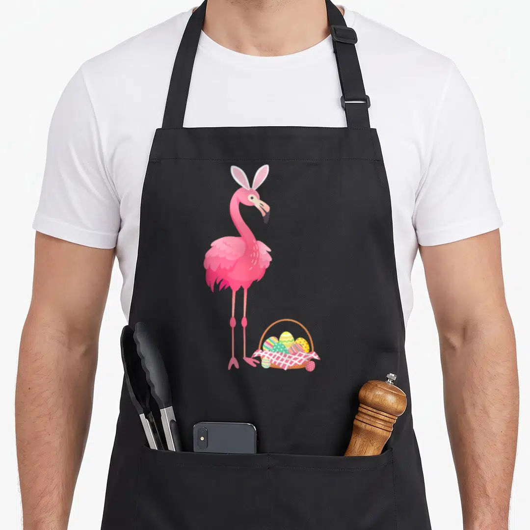 Flamingo basket apron - Pink flamingo wearing white bunny ears standing next to a woven basket filled with decorated — shown on a black cotton canvas apron against a white background
