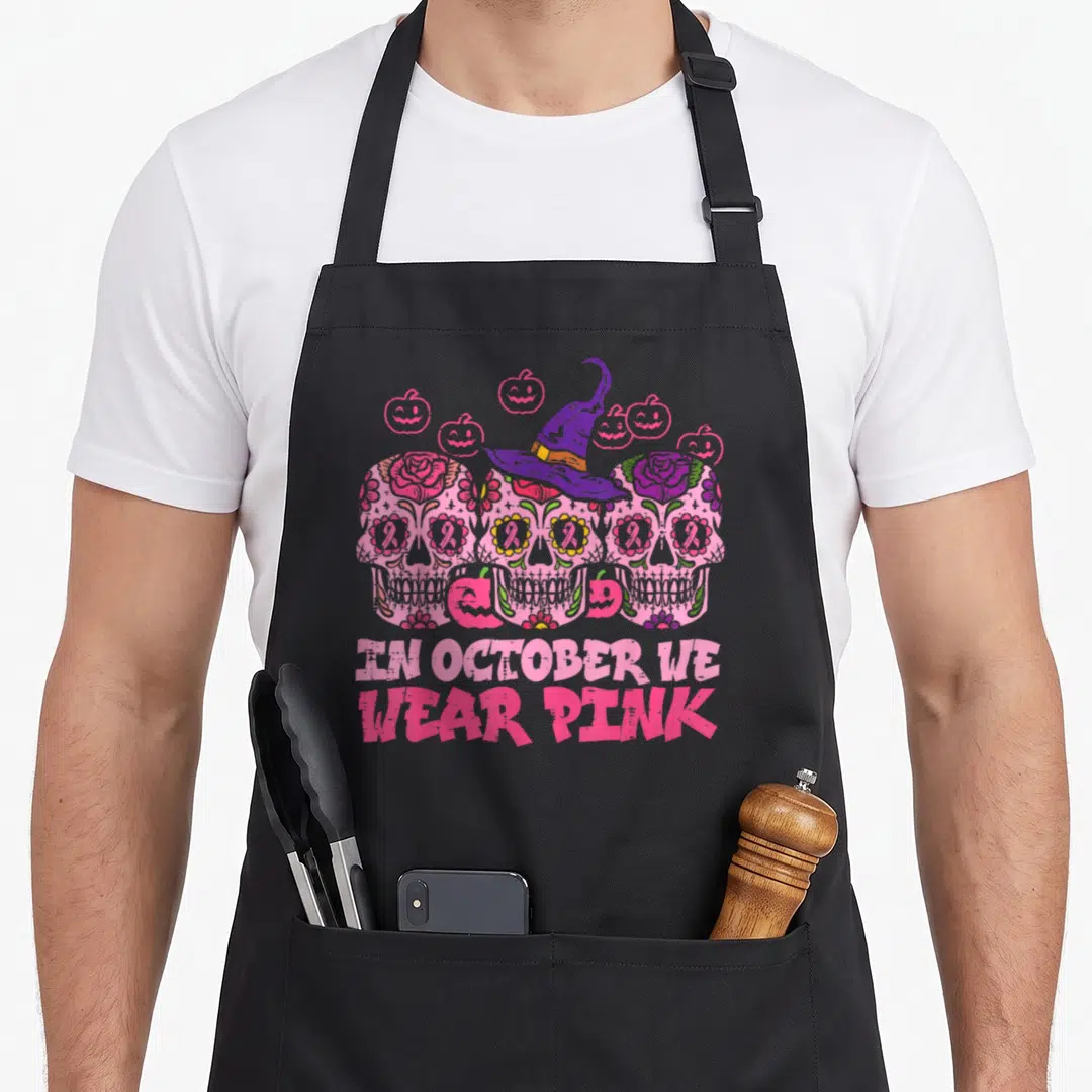 October sugar skull apron - Three pink sugar skulls with pink ribbon eyes, purple witch hat, orange pumpkins — shown on a black cotton canvas apron against a white background