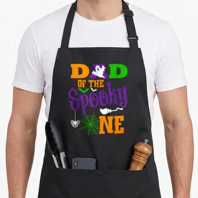Family funny halloween apron - Dad of the Spooky One text with ghost bat spider and web in orange purple green and white — shown on a black cotton canvas apron against a white background
