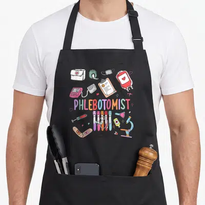 Phleb nurse apron - Phlebotomist text with cartoon syringes, blood bag, and medical puns in pink blue red yellow — shown on a black cotton canvas apron against a white background