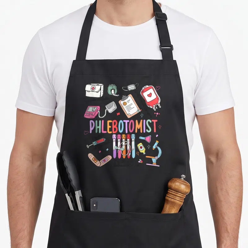 Phleb nurse apron - Phlebotomist text with cartoon syringes, blood bag, and medical puns in pink blue red yellow — shown on a black cotton canvas apron against a white background