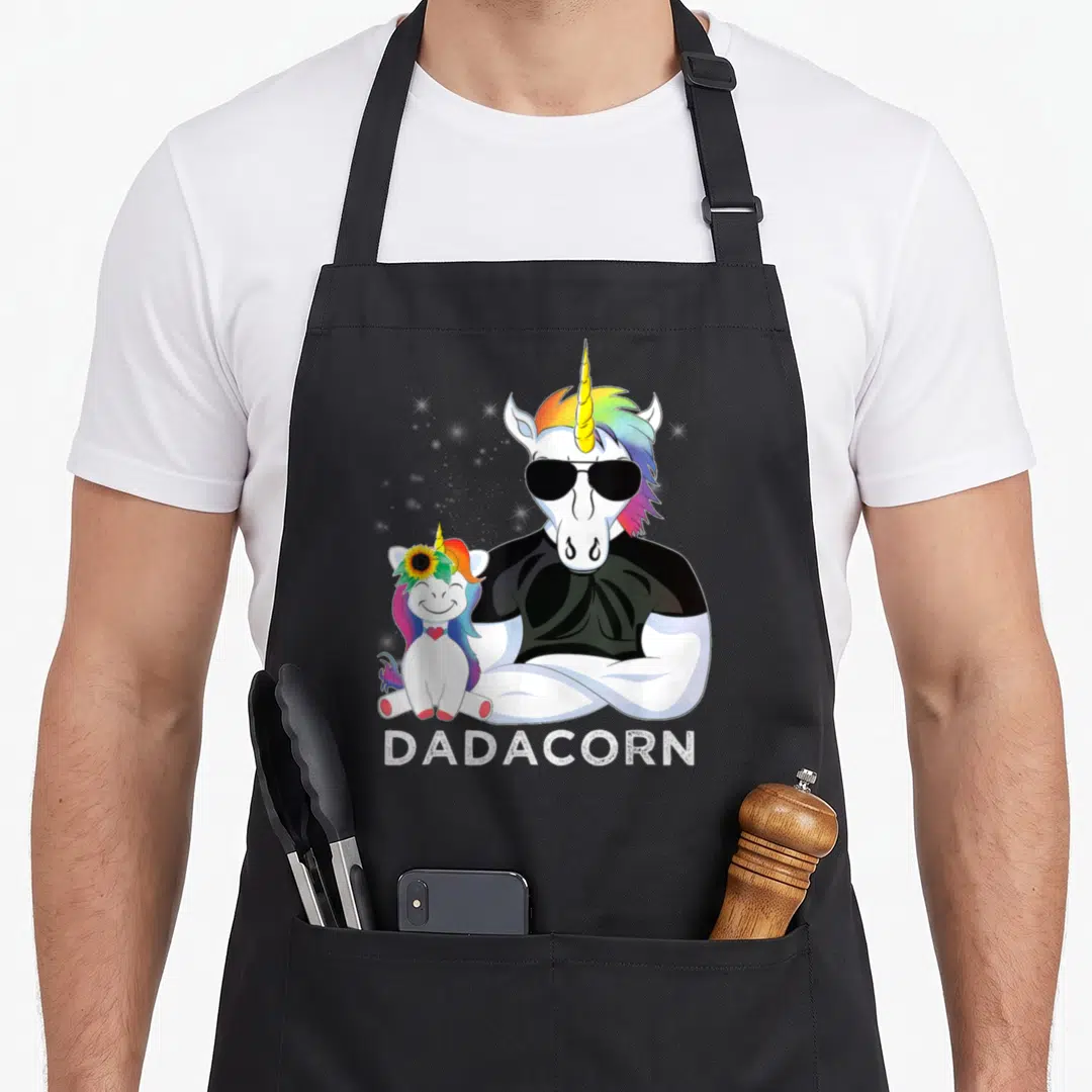 Dadacorn muscle apron - Cartoon unicorn in sunglasses and black shirt stands behind baby unicorn with sunflower — shown on a black cotton canvas apron against a white background