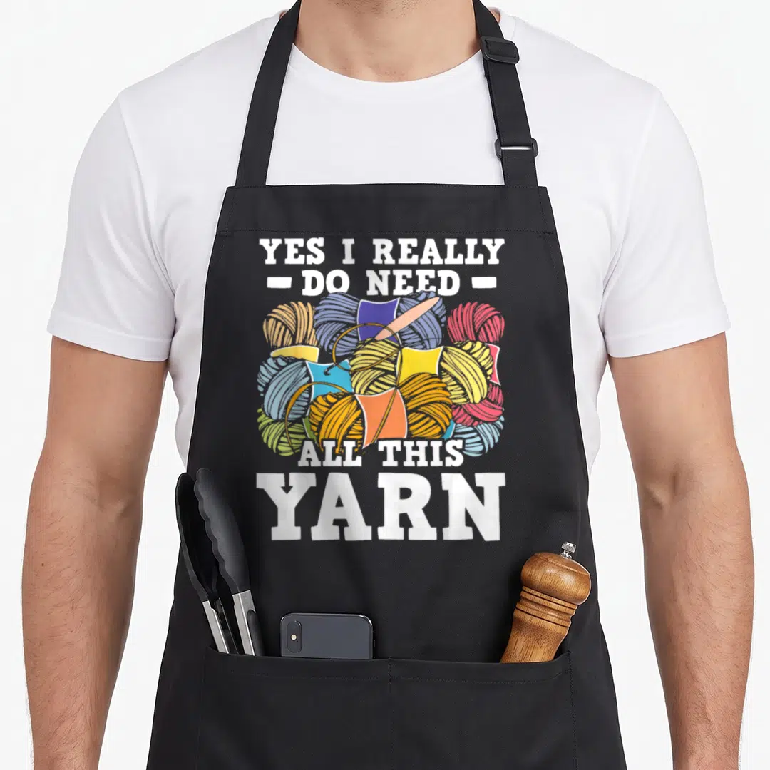 Yarn do i need an apron - Cartoon yarn balls in blue yellow red orange white with pink needle and bold text — shown on a black cotton canvas apron against a white background