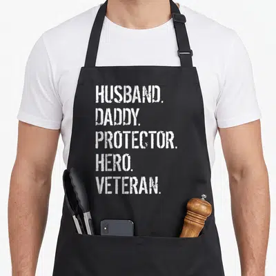 Father best husband apron - White stenciled text reading HUSBAND DADDY PROTECTOR HERO VETERAN stacked vertically — shown on a black cotton canvas apron against a white background