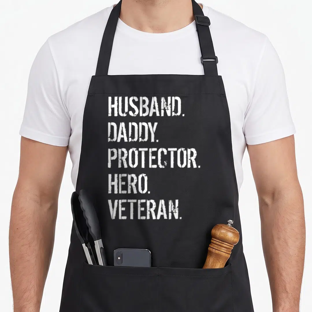Father best husband apron - White stenciled text reading HUSBAND DADDY PROTECTOR HERO VETERAN stacked vertically — shown on a black cotton canvas apron against a white background