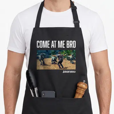 Come jurassic world apron - Man facing three dinosaurs under white Come At Me Bro text on black bib apron — shown on a black cotton canvas apron against a white background