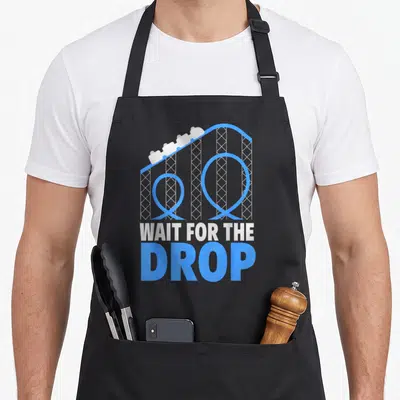 Park roller apron - Blue and grey roller coaster track with cars above stacked text WAIT FOR THE DROP — shown on a black cotton canvas apron against a white background